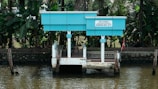 A small, blue and white jetty is situated on the edge of a river, surrounded by lush green tropical foliage. The structure has a board that reads 'Govt. of Kerala Grameena Boat Jetty'. It's a quiet and serene location, ideal for boarding small boats.