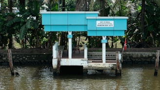 A small, blue and white jetty is situated on the edge of a river, surrounded by lush green tropical foliage. The structure has a board that reads 'Govt. of Kerala Grameena Boat Jetty'. It's a quiet and serene location, ideal for boarding small boats.