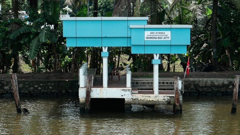 A small, blue and white jetty is situated on the edge of a river, surrounded by lush green tropical foliage. The structure has a board that reads 'Govt. of Kerala Grameena Boat Jetty'. It's a quiet and serene location, ideal for boarding small boats.
