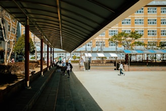A school courtyard with a covered walkway on the left side leading towards the center. Several students are walking and sitting in the area, with some greenery and trees in the background. The building in the background has multiple windows, suggesting a multi-story school structure.