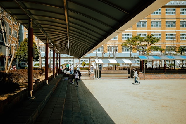 A school courtyard with a covered walkway on the left side leading towards the center. Several students are walking and sitting in the area, with some greenery and trees in the background. The building in the background has multiple windows, suggesting a multi-story school structure.