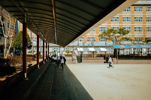 A school courtyard with a covered walkway on the left side leading towards the center. Several students are walking and sitting in the area, with some greenery and trees in the background. The building in the background has multiple windows, suggesting a multi-story school structure.