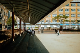 A school courtyard with a covered walkway on the left side leading towards the center. Several students are walking and sitting in the area, with some greenery and trees in the background. The building in the background has multiple windows, suggesting a multi-story school structure.