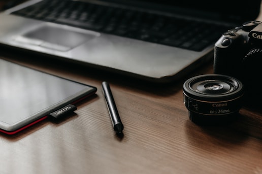 A workspace scene with a tablet, stylus, camera lens, and a DSLR camera placed on a wooden table surface. In the background, an open laptop's keyboard and touchpad are partially visible, creating a setup suited for digital creativity or photography work.