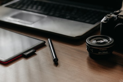 A workspace scene with a tablet, stylus, camera lens, and a DSLR camera placed on a wooden table surface. In the background, an open laptop's keyboard and touchpad are partially visible, creating a setup suited for digital creativity or photography work.