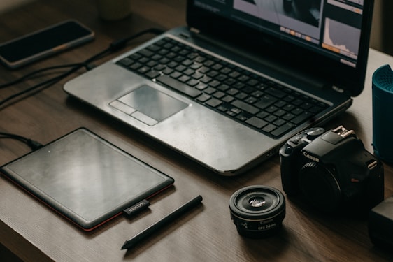 A wooden table with a laptop displaying photo editing software, alongside a graphics tablet, pen, DSLR camera, camera lens, and a smartphone.