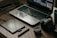 A wooden table with a laptop displaying photo editing software, alongside a graphics tablet, pen, DSLR camera, camera lens, and a smartphone.