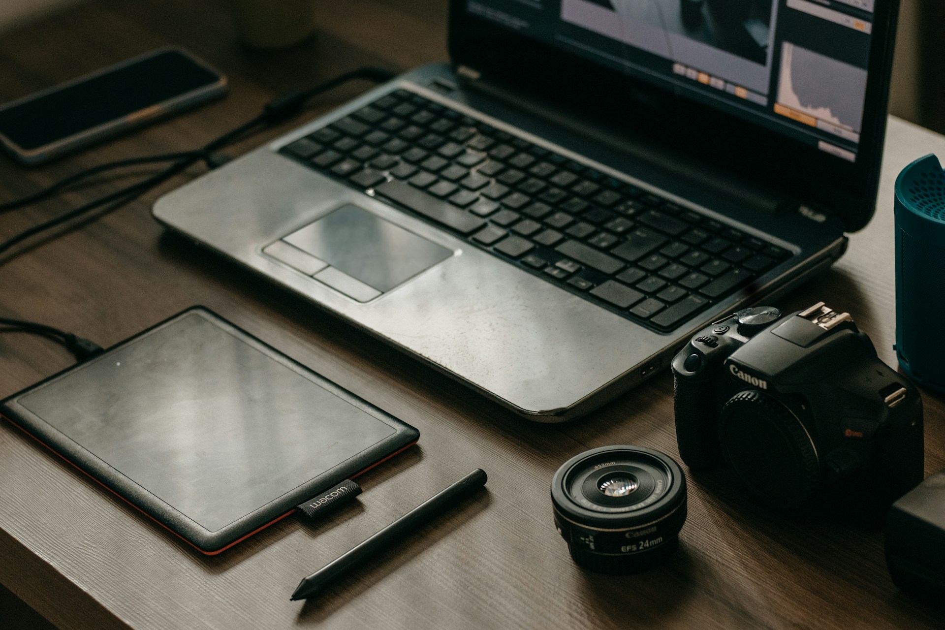 A wooden table with a laptop displaying photo editing software, alongside a graphics tablet, pen, DSLR camera, camera lens, and a smartphone.