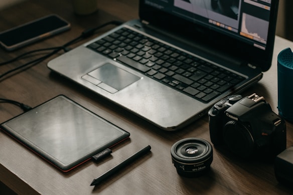 A wooden table with a laptop displaying photo editing software, alongside a graphics tablet, pen, DSLR camera, camera lens, and a smartphone.