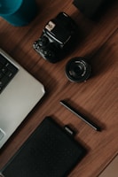 A top-down view of a wooden desk featuring various gadgets, including a digital camera, a camera lens, a graphic tablet with a stylus, a part of a laptop keyboard, and a blue cylindrical speaker.