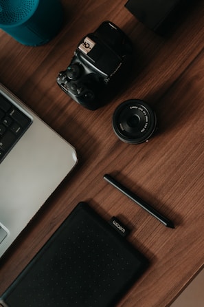 A top-down view of a wooden desk featuring various gadgets, including a digital camera, a camera lens, a graphic tablet with a stylus, a part of a laptop keyboard, and a blue cylindrical speaker.
