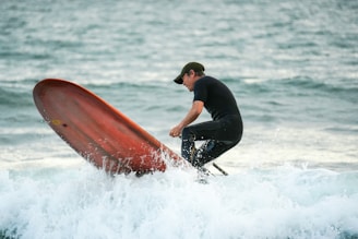 A person wearing a black wetsuit and dark cap is surfing on a body of water. The individual appears to be struggling with balancing on the surfboard, which is tipped upwards against the waves. The background consists of wavy ocean water with a slight blur.