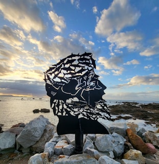 A silhouette sculpture made of metal depicting the face of a man, comprised of various marine animals like sharks and fish, stands against a coastal backdrop. The sky is filled with soft clouds reflecting the light of a setting or rising sun, while rocky formations line the shore.