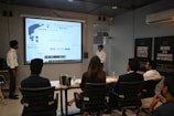 A presentation takes place in a modern conference room with a group of professionals seated, and two presenters standing near a projected screen. The screen displays a web page with the McAfee Security Scan Plus showing. The atmosphere is formal and focused, as attendees appear to be discussing or reviewing the content.