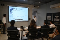 A presentation takes place in a modern conference room with a group of professionals seated, and two presenters standing near a projected screen. The screen displays a web page with the McAfee Security Scan Plus showing. The atmosphere is formal and focused, as attendees appear to be discussing or reviewing the content.