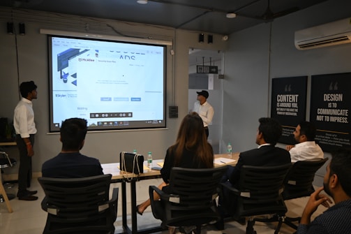 A presentation takes place in a modern conference room with a group of professionals seated, and two presenters standing near a projected screen. The screen displays a web page with the McAfee Security Scan Plus showing. The atmosphere is formal and focused, as attendees appear to be discussing or reviewing the content.