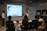 A presentation takes place in a modern conference room with a group of professionals seated, and two presenters standing near a projected screen. The screen displays a web page with the McAfee Security Scan Plus showing. The atmosphere is formal and focused, as attendees appear to be discussing or reviewing the content.