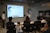 A presentation takes place in a modern conference room with a group of professionals seated, and two presenters standing near a projected screen. The screen displays a web page with the McAfee Security Scan Plus showing. The atmosphere is formal and focused, as attendees appear to be discussing or reviewing the content.