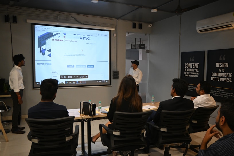 A presentation takes place in a modern conference room with a group of professionals seated, and two presenters standing near a projected screen. The screen displays a web page with the McAfee Security Scan Plus showing. The atmosphere is formal and focused, as attendees appear to be discussing or reviewing the content.