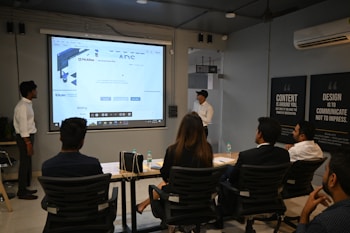 A presentation takes place in a modern conference room with a group of professionals seated, and two presenters standing near a projected screen. The screen displays a web page with the McAfee Security Scan Plus showing. The atmosphere is formal and focused, as attendees appear to be discussing or reviewing the content.