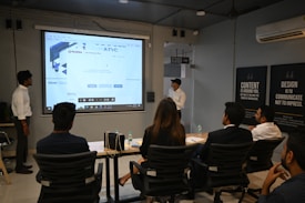 A presentation takes place in a modern conference room with a group of professionals seated, and two presenters standing near a projected screen. The screen displays a web page with the McAfee Security Scan Plus showing. The atmosphere is formal and focused, as attendees appear to be discussing or reviewing the content.