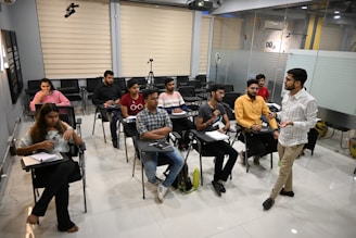 A group of people are in a classroom setting. Some are seated at desks with notebooks and pens, while one person appears to be teaching or giving a presentation. The classroom has a modern design with glass partitions and various electronic equipment, such as a tripod and a ring light.
