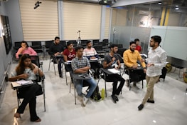A group of people are in a classroom setting. Some are seated at desks with notebooks and pens, while one person appears to be teaching or giving a presentation. The classroom has a modern design with glass partitions and various electronic equipment, such as a tripod and a ring light.