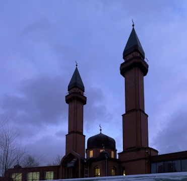 A mosque featuring two tall brown brick minarets with green pointed roofs and crescent ornaments. The central dome is illuminated from inside, creating a contrast with the cloudy evening sky.