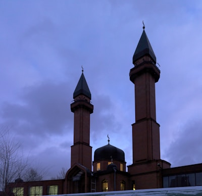 A mosque featuring two tall brown brick minarets with green pointed roofs and crescent ornaments. The central dome is illuminated from inside, creating a contrast with the cloudy evening sky.