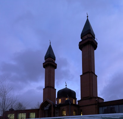 A mosque featuring two tall brown brick minarets with green pointed roofs and crescent ornaments. The central dome is illuminated from inside, creating a contrast with the cloudy evening sky.