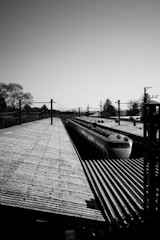 A black and white image of a train station platform, featuring a high-speed train. The platform has a textured surface with shadows casting across it. The scene is framed by overhead power lines and trees in the background, under a clear sky.