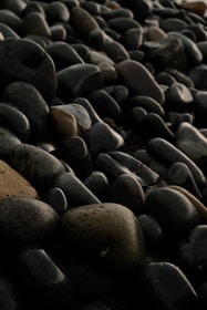 A close-up of a collection of smooth, dark-colored stones. The stones vary in size but share similar rounded shapes. Some of the stones have a slightly greenish hue while others are more brown or black. The lighting creates a subtle play of shadows and highlights on the surfaces of the stones.