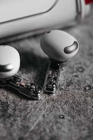 Two wireless earbuds with a sleek and modern design are lying on a textured gray surface. The earbuds feature a transparent section revealing internal components, adding a futuristic element to their appearance.