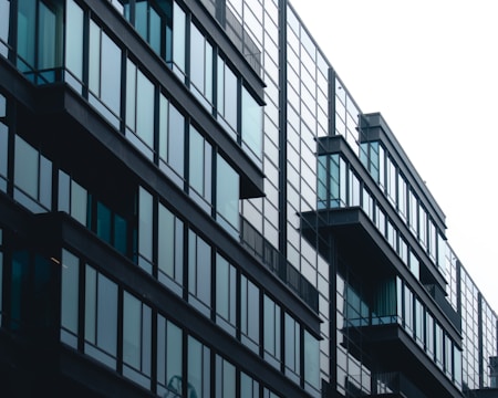 A modern building with a sleek, minimalist design featuring large glass windows and balconies extending from each floor. The structure appears symmetrical, with reflective surfaces that suggest a contemporary architectural style.