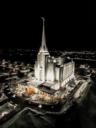 A beautifully illuminated temple stands in the center of a snow-covered landscape at night. The architecture features tall spires and intricate details, with soft yellow lighting accentuating its structure. The surrounding area is serene, with snow on the ground and bare trees framing the scene. In the background, lights from a distant city are visible.