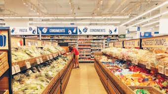 A well-lit supermarket aisle featuring a section of produce on the left and a variety of packaged foods on the right. The ceiling is equipped with bright fluorescent lights. Two people are standing in the aisle, selecting items. Signs above indicate sections for pasta and condiments.