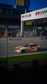 A racing car with a red and yellow livery speeds past on a racing track. The track is lined with metal fencing and banners advertising a performance exhaust brand. A digital clock displays the time above the track, and a scoreboard is visible in the background, along with the hashtag #AUSTRIANGP.