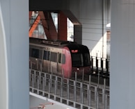 A modern train is situated on elevated tracks surrounded by industrial architecture. The train has a sleek design with a shiny metallic body, red accents, and digital signage on the front. The setting includes large structural beams and wires, suggesting an urban transit system.