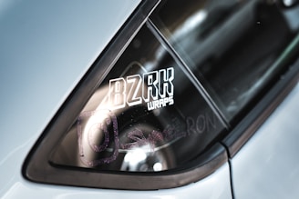 The image features a close-up of a car window with a vinyl wrap logo. The text 'BZRK WRAPS' is prominently displayed in bold and stylized lettering. There are also faint reflections and another symbol resembling an Instagram logo nearby.