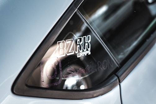 The image features a close-up of a car window with a vinyl wrap logo. The text 'BZRK WRAPS' is prominently displayed in bold and stylized lettering. There are also faint reflections and another symbol resembling an Instagram logo nearby.