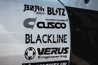 Multiple brand logos are displayed on a white surface, featuring various automotive and engineering companies such as Blitz, Cusco, Blackline, and Verus Engineering. The text is printed in bold black font, each with distinctive logo designs.