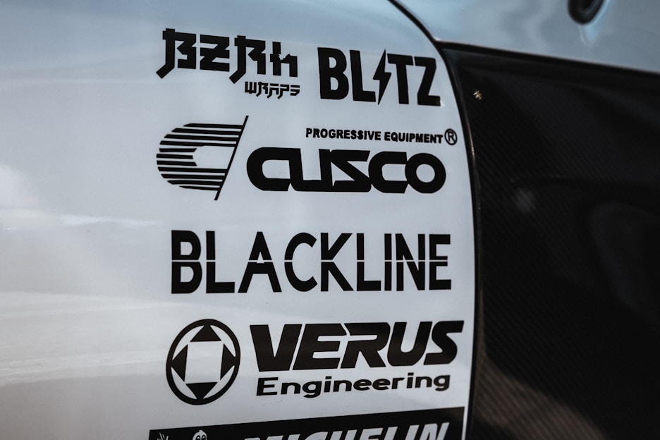 Multiple brand logos are displayed on a white surface, featuring various automotive and engineering companies such as Blitz, Cusco, Blackline, and Verus Engineering. The text is printed in bold black font, each with distinctive logo designs.