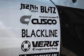 Multiple brand logos are displayed on a white surface, featuring various automotive and engineering companies such as Blitz, Cusco, Blackline, and Verus Engineering. The text is printed in bold black font, each with distinctive logo designs.