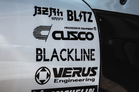 Multiple brand logos are displayed on a white surface, featuring various automotive and engineering companies such as Blitz, Cusco, Blackline, and Verus Engineering. The text is printed in bold black font, each with distinctive logo designs.
