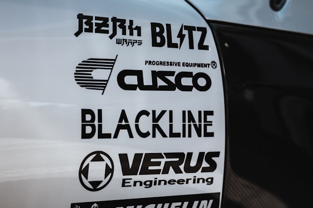 Multiple brand logos are displayed on a white surface, featuring various automotive and engineering companies such as Blitz, Cusco, Blackline, and Verus Engineering. The text is printed in bold black font, each with distinctive logo designs.