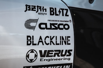 Multiple brand logos are displayed on a white surface, featuring various automotive and engineering companies such as Blitz, Cusco, Blackline, and Verus Engineering. The text is printed in bold black font, each with distinctive logo designs.