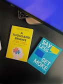 Two books placed on a desk with part of a keyboard visible. One book has a yellow cover titled 'A Thousand Brains' by Jeff Hawkins, featuring an illustration of a human head with puzzle pieces. The other book has a turquoise cover titled 'Say Less, Get More' by Fotini Iconomopoulos, with speech bubbles on the cover. The setting appears to be a workspace.