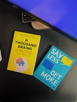 Two books placed on a desk with part of a keyboard visible. One book has a yellow cover titled 'A Thousand Brains' by Jeff Hawkins, featuring an illustration of a human head with puzzle pieces. The other book has a turquoise cover titled 'Say Less, Get More' by Fotini Iconomopoulos, with speech bubbles on the cover. The setting appears to be a workspace.