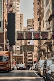 A busy urban street lined with tall buildings and signs in Chinese, featuring a variety of parked cars and a moving truck. Pedestrians are seen walking along the sidewalk, while traffic is visible in the background under the narrow strip of sky.