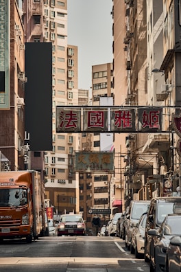 A busy urban street lined with tall buildings and signs in Chinese, featuring a variety of parked cars and a moving truck. Pedestrians are seen walking along the sidewalk, while traffic is visible in the background under the narrow strip of sky.